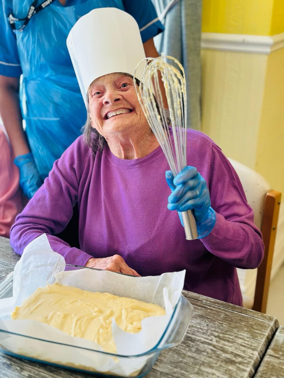 A smiling resident in a chef's hat holding a whisk over a tray of cake mix